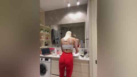 Media: Video of a slender, blonde woman with medium build, wearing a white sports bra and red leggings, looking into a large mirror in a modern, minimalist bathroom with white cabinets and a washer-dryer.