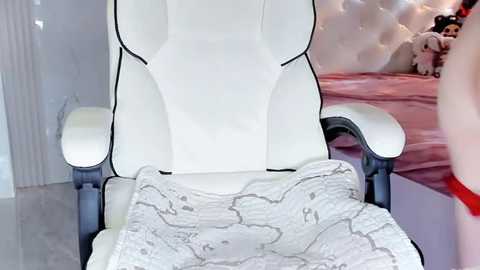 Media: A video of a plush, white gaming chair with a quilted blanket featuring a map pattern, placed in a cozy, pink bedroom with a tufted headboard and stuffed animals.