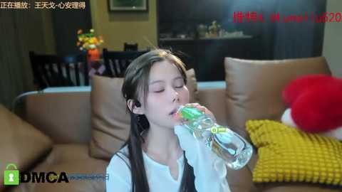 Media: Video of a young Asian girl with long brown hair, wearing a white shirt, drinking from a clear plastic bottle in a modern living room with beige furniture, a yellow throw, and a red plush toy.