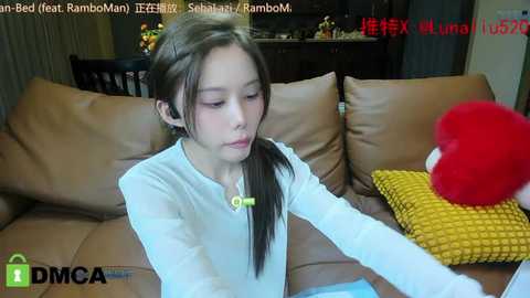 Media: Video of a young Asian girl with long brown hair, wearing a white long-sleeve shirt, sitting on a brown leather couch with a yellow knitted pillow. Background shows a dimly lit room with a dark wooden cabinet and a \"DMCA\" watermark.