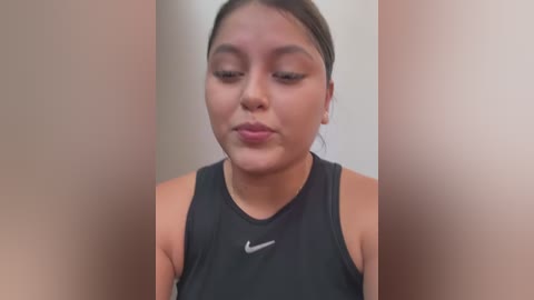 Media: Video of a young woman with medium skin tone, medium build, and dark hair tied back. She wears a black Nike sports top. Background is blurred, focusing on her face.