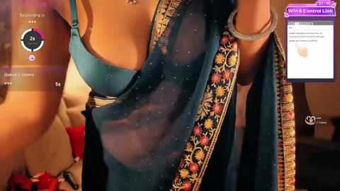 Media: A video of a woman in a teal saree with gold embroidery, wearing a matching blue bra, seen from the side, in a dimly lit room.