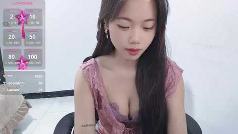 Media: Video of an Asian woman with long black hair, wearing a pink lace dress, seated indoors. Background includes a calendar and a white wall.