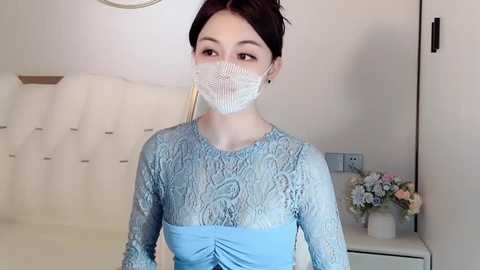 Media: Video of a young Asian woman with fair skin and dark hair in a bun, wearing a blue lace dress and white face mask, standing in a modern, minimalist bedroom with white walls, a bed, and floral arrangement.