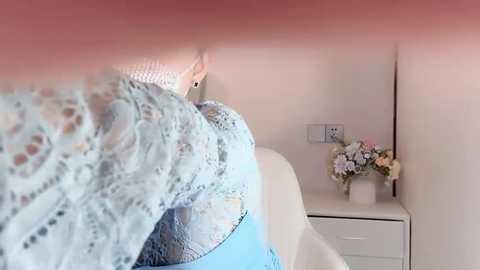 Video of a woman's torso in a light blue lace top, wearing blue jeans, standing in a corner with a white dresser and floral arrangement.