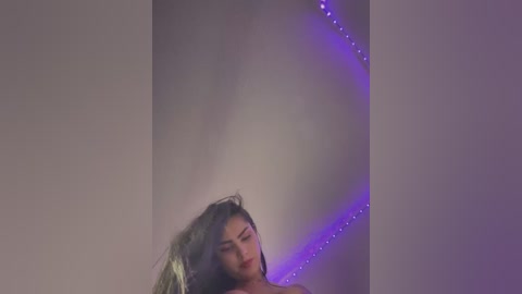 Video of a woman with long, dark hair and fair skin, standing in a dimly lit room with a purple light strip behind her.