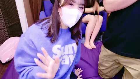 Media: A video of an East Asian woman with straight, shoulder-length brown hair and a white surgical mask, wearing a blue \"Sunway\" sweatshirt, seated on a purple couch. A man stands beside her, partially visible.