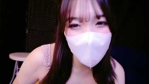 Media: A video of an Asian woman with long black hair and fair skin, wearing a white face mask and a light-colored top, against a dimly lit background.