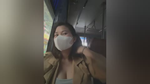 Media: A video of an East Asian woman with medium skin tone, wearing a white face mask and an unbuttoned tan jacket, revealing a low-cut top, in a dimly lit subway car.