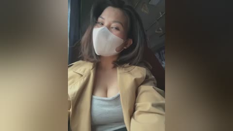 Media: Video of a woman with medium skin tone, short dark hair, wearing a beige jacket over a white tank top, a white mask, and a light blue face shield. She has a neutral expression.