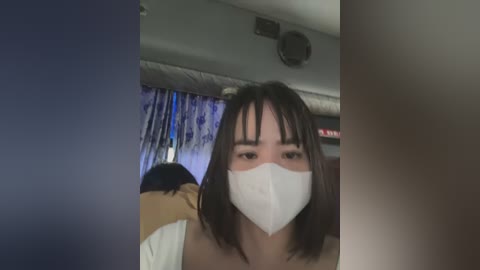 Media: A video of a young woman with straight, dark hair and bangs wearing a white face mask and a yellow top. She is indoors, possibly on a plane, with a blue and white patterned curtain and overhead lighting in the background.