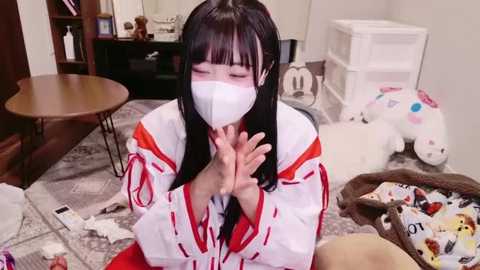 Media: Video of an Asian woman in traditional Japanese kimono, wearing a white face mask, hands clasped, kneeling on a gray carpeted floor. Background features a wooden table, bookshelf, stuffed toys, and scattered toys.