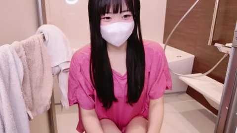 Media: A video of an Asian woman with long black hair, wearing a white mask, pink T-shirt, and sitting on a white tiled floor in a modern bathroom with beige walls and a white towel hanging.