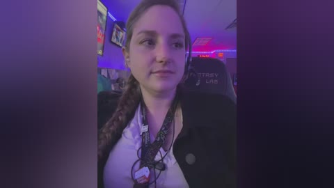 Media: Video of a young woman with long brown hair in braids, wearing a black jacket over a white shirt, sitting in a dark gaming chair. The room is dimly lit with purple and red lighting, featuring various posters and gaming equipment.