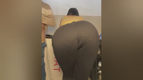 Media: A video of a woman with a large, round buttocks, dressed in tight, dark gray leggings, leaning over a table, partially obscured by a beige curtain.