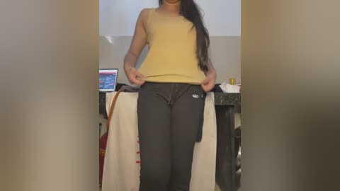 Media: Video of a woman with long black hair, wearing a mustard tank top and dark jeans, standing in a cluttered room with a computer and a white towel.