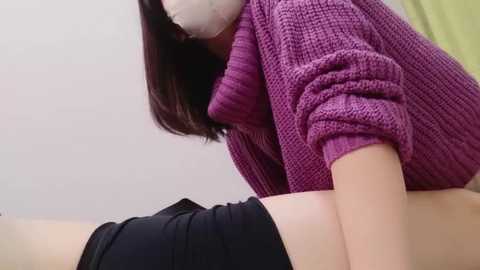 Media: Video of a person with shoulder-length dark hair, wearing a purple knit sweater and black shorts, lying on a white surface. The background is plain white, and a yellow object is partially visible.