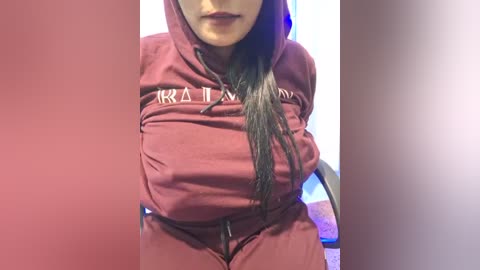 Media: Video of a woman with long black hair, wearing a maroon hoodie with \"R.A.M.\" in white, her breasts prominently visible through the fabric, seated in a chair against a blurred background.