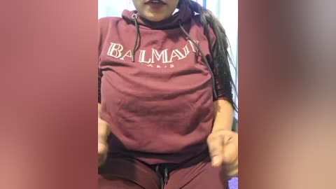 Media: Video of a woman wearing a maroon \"Balmain Paris\" sweatshirt with a hood, revealing her ample breasts and belly, with a blurred background.