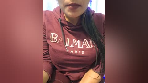 Media: Video of a woman with light skin and long black hair, wearing a maroon Balmain Paris hoodie, partially cropped face visible.