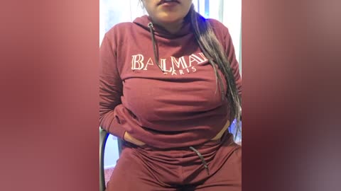 Media: A video of a plus-sized woman in a red Balmain Paris hoodie and matching sweatpants, with long braids and hands in pockets. Background features vertical blue and white stripes.