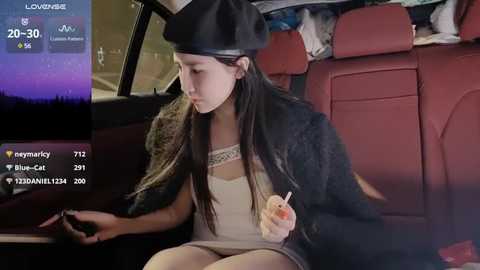 Media: Video of a young East Asian woman in a car, wearing a black beret, beige dress, and fur jacket, texting, holding a drink, with a smartphone in her hand.