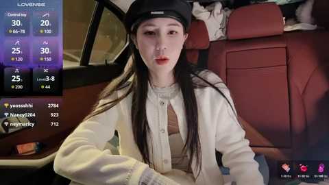 Media: A video of an East Asian woman with long black hair, fair skin, and red lipstick, wearing a black cap, white sweater, and beige jacket, sitting in a red leather car seat. The background shows car controls and a map.