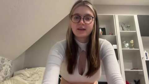 Media: Video of a young Caucasian woman with long brown hair, wearing glasses, a white ribbed top with a deep V-neck, standing in a modern, minimalist bedroom.