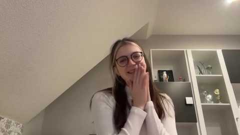 Media: A video of a young woman with glasses, long brown hair, wearing a white sweater, smiling and covering her mouth. She stands in a modern, minimalist bedroom with beige walls, white shelves, and decorative items.