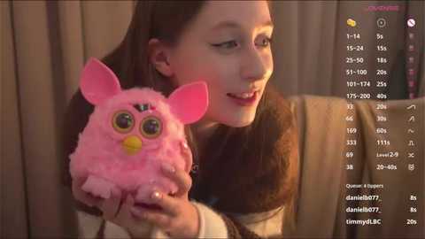 Video of a smiling woman with light skin and brown hair, holding a pink, fluffy, googly-eyed plush toy, against a beige curtain background.