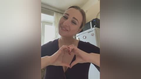 Media: Video of a smiling Caucasian woman with light skin and brown hair in a dark top, forming a heart shape with her hands in a cozy kitchen with a white microwave and cream-colored curtains.