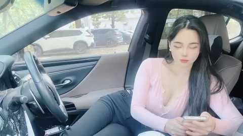 Media: Video of an attractive East Asian woman with long black hair, wearing a low-cut pink top, sitting in a car, texting on her phone.