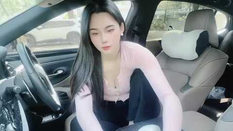 Media: Video of an Asian woman with long black hair, wearing a pink top, sitting in a car with beige seats and a steering wheel.