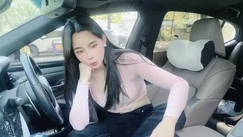 Media: Video of an Asian woman with long black hair, wearing a pink top, leaning on the car seat, looking tired. Car interior with beige seats and a dashboard.