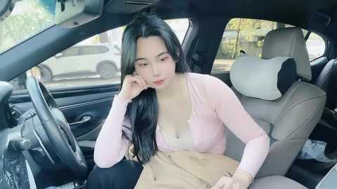 Media: Video of an East Asian woman with long black hair, fair skin, and a slender physique, wearing a light pink cardigan and beige pants, sitting in a car with beige leather seats, looking tired and contemplative.