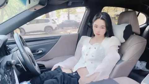 Media: Video of a young Asian woman with long black hair, dressed in a white blouse and black pants, sitting in a modern, beige-carpeted car interior.