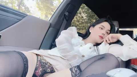 A video of a young woman with fair skin and long black hair, wearing a white blouse, black lace panties, and thigh-high stockings, reclining seductively in a car.