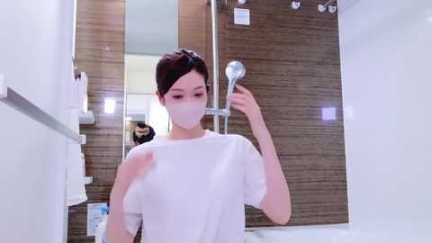 Media: Video of a young East Asian woman with light skin, brown hair tied back, wearing a white mask and shirt, taking a selfie in a modern, clean bathroom with wooden wall panels and white fixtures.