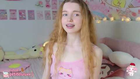 Media: Video of a young, fair-skinned woman with long, wavy blonde hair, wearing a pink tank top with a cartoon cat design. She smiles gently, surrounded by plush toys in a pastel-themed bedroom.