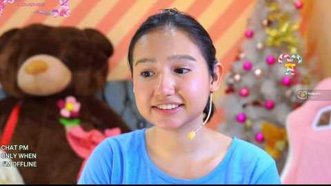 Media: A video of a smiling young woman with dark hair, wearing a blue shirt, sitting in a festive room with a Christmas tree and teddy bear in the background.