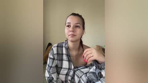 Media: Video of a young woman with light skin, brown hair tied back, wearing a black and white plaid shirt. She sits in a chair with a neutral background.