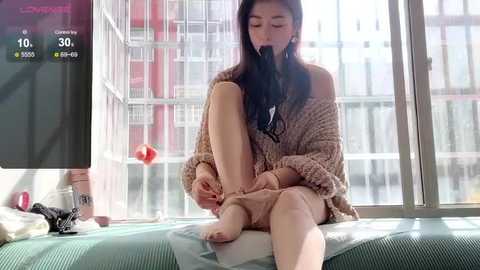 Video of an Asian woman with long black hair, wearing a beige off-shoulder sweater, sitting on a bed, tying her beige socks. Background shows a modern apartment with a large window and city view.