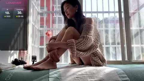 Media: Video of an Asian woman in a knitted off-shoulder sweater, sitting on a futon, barefoot, gazing out a large window at a modern urban skyline.