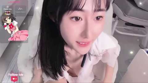 Media: Video of a young Asian woman with black hair, wearing a white blouse, in a modern, sterile room. The image appears digitally altered with a \"Follow Me\" watermark and a small anime character in the corner.