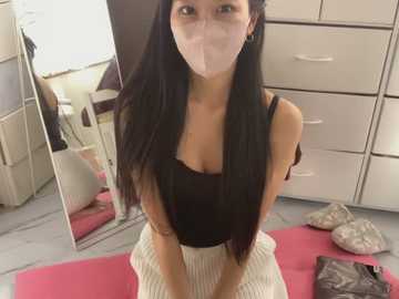 Media: Video of a young Asian woman with long black hair, wearing a black tank top and white skirt, kneeling on a pink yoga mat, in a bedroom with beige furniture and mirrors.