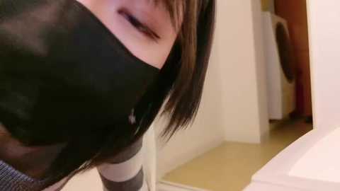 Media: Video of a woman with straight, shoulder-length brown hair, wearing a black surgical mask, smiling. Background shows a beige tiled floor and a white washing machine.