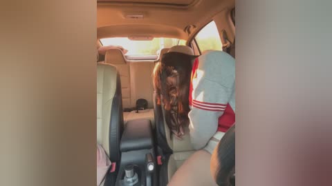 Media: Video of a woman with long, dark hair in a grey shirt and red shorts, seated in the backseat of a car, with beige seats and a sunlit view outside.