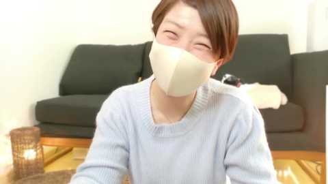 Media: Video of an Asian woman with short brown hair, wearing a light blue sweater and a white mask, sitting on a wooden floor with a black sofa and a brown lamp in the background.