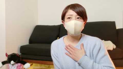 Media: A video of an East Asian woman with short brown hair, wearing a light gray sweater and white surgical mask, standing indoors next to a black sofa, holding her chest.