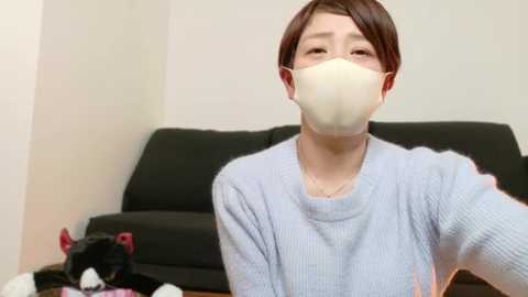 Media: A video of an East Asian person with short brown hair, wearing a light gray sweater, beige face mask, and headphones, sitting on a black sofa in a minimalist living room.